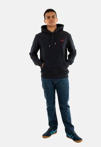 Sudadera de un oscuro color azul marino con un bolsillo frontal y cordones. Presenta un logo rojo en el pecho, combinada con jeans azules y zapatillas negras con detalles azules.