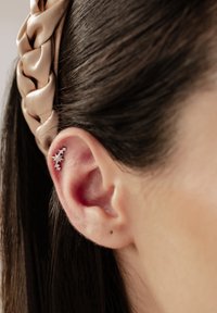 Pendiente de oído plateado con acentos de estrellas y piedras preciosas, sobre un fondo de cabello liso y oscuro y una diadema trenzada beige.