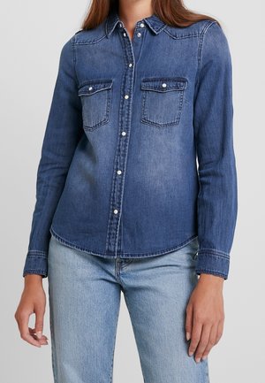 Kobieta w niebieskiej koszuli dżinsowej z zapięciem na guziki, posiadającej dwie kieszenie na piersi oraz jasnoniebieskie dżinsy, na tle jednolitego szarego tła.