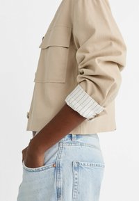 Beige cropped jacket with two chest pockets and rolled-up striped white cuffs. Worn over light blue denim shorts.