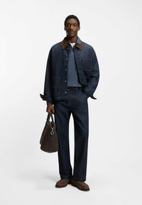 Kurtka dżinsowa z brązowym kołnierzem, niebieska koszulka polo i dopasowane dżinsy. Brązowa torba w ręku, brązowe buty, proste nogawki, bez widocznych wzorów.