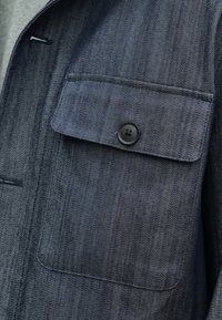 Sötétkék farmerdzseki zsebe, amelyet egyetlen fekete gombbal rögzített zsebfül fedi, részletesen látható a szövet textúrája és a varrás.