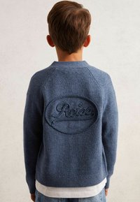 Kék kötött pulóver, raglán ujjakkal, emelt hímzett logóval a hátsó részén és kerek nyakkivágással. Fehér ing fölé rétegezve.