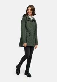 Zielona wodoodporna kurtka z kapturem, regulowanym ściągaczem w pasie, dwoma przednimi kieszeniami i zapięciem na zamek. Połączona z czarnymi dżinsami i butami.