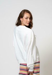 Fehér, oversized kapucnis pulóver kerek nyakkivágással és ejtett vállakkal, amit több színben csíkos kötött nadrággal párosítottak. Egyszerű háttér.