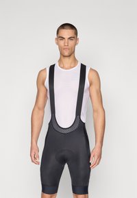 Svarte sykkelshorts med mesh-stropper, med polstret sete, glatt stoff og tettsittende passform. Brukt over en hvit mesh singlet.