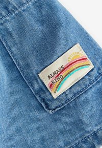 Patch sur tissu en denim bleu avec un motif coloré d’arc-en-ciel et de soleil, affichant le texte « TOUJOURS GENTIL ».