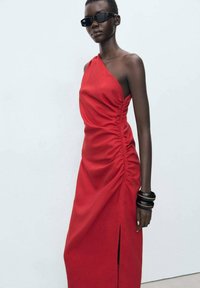Robe rouge à une épaule avec des détails froncés et une fente sur le côté, fabriquée en tissu lisse ; associée à des bracelets noirs et des lunettes de soleil.