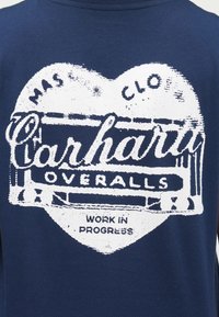 Marineblå t-shirt med en slidt hvid grafik, der viser teksten "Carhartt Overalls" og "Work in Progress" i et hjerteformet design.