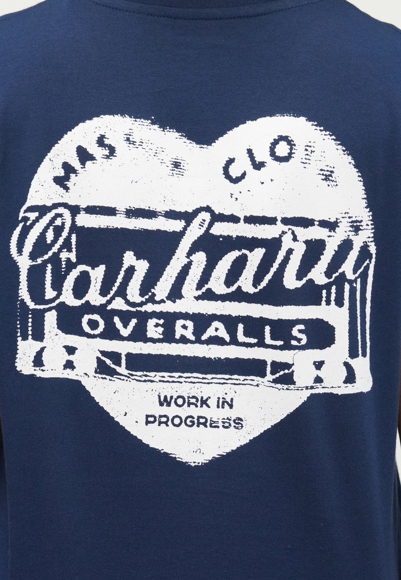 Marineblaue T-Shirt mit einem abgenutzten weißen Grafikdesign, das "Carhartt Overalls" und "Work in Progress" in einer herzförmigen Anordnung zeigt.