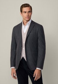 Uomo che indossa un blazer grigio scuro sopra un cardigan beige e una camicia bianca con bottoni, in piedi contro uno sfondo grigio chiaro uniforme.