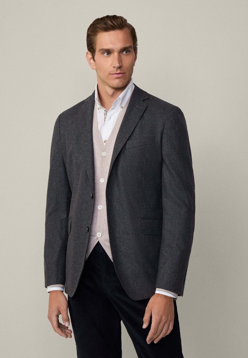 Uomo che indossa un blazer grigio scuro sopra un cardigan beige e una camicia bianca con bottoni, in piedi contro uno sfondo grigio chiaro uniforme.