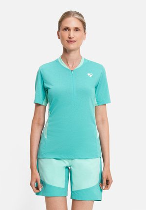 Frau trägt helltürkisfarbenes, kurzärmeliges Oberteil mit Reißverschluss und passende Shorts mit Farbfeldern, stehend vor weißem Hintergrund.