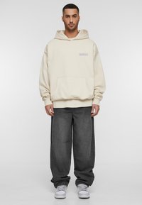 Creme hoodie med frontlomme, afslappet pasform, broderet tekst i lys lilla; parret med løst siddende mørkegrå jeans og hvide sneakers.