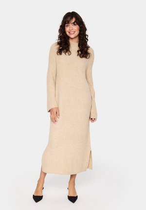 Knit beige sweater dress with long sleeves, high neckline, side slit, and ribbed texture. Paired with black pointed flats.