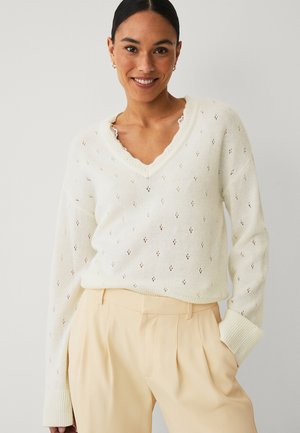 Frau lächelnd, trägt einen weißen, strukturierten V-Ausschnitt-Pullover und beige, hoch taillierte Hosen, steht mit einer Hand in der Tasche vor einem schlichten Hintergrund.