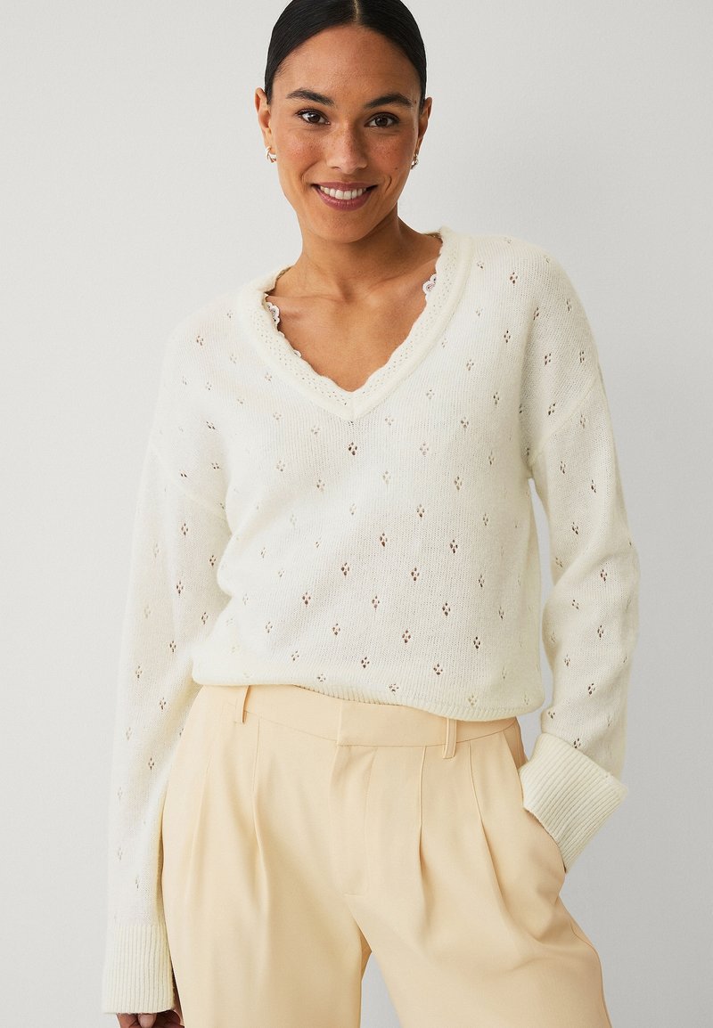 Žena se usmívá, nosí bílý texturovaný svetr s výstřihem do V a béžové kalhoty s vysokým pasem, stojí s jednou rukou v kapse na jednobarevném pozadí.