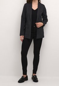 Zwarte pinstripe blazer met notch-klepels, twee voorzakken en een lichtgewicht stof, gecombineerd met zwarte slim-fit jeans en instappers.