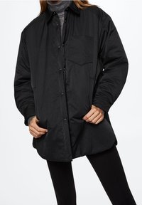 Zwarte, oversized overhemdjas van gladde, lichtgewicht stof. Bevat een borstzak en knoopsluiting, met een relaxte pasvorm.