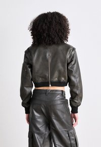 Krótka czarna skórzana kurtka bomberka z prążkowanymi mankietami, zamkiem błyskawicznym i gładką fakturą. Noszona z dopasowanymi skórzanymi spodniami.