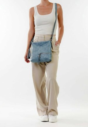 Borsa a tracolla in pelle blu con superficie texture, dotata di una tasca con zip frontale e un accento a nappina, indossata con pantaloni beige a gamba larga.