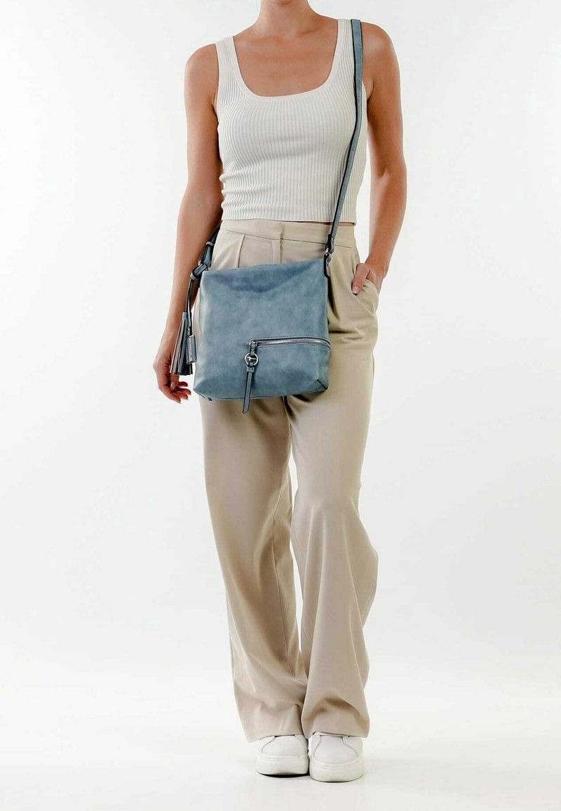 Borsa a tracolla in pelle blu con superficie texture, dotata di una tasca con zip frontale e un accento a nappina, indossata con pantaloni beige a gamba larga.