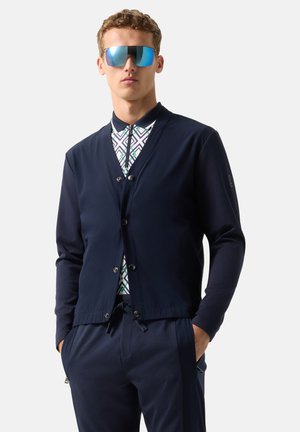 Mand iført mørkeblå jakke over mønstret lynlås-skjorte, mørkeblå bukser og reflekterende blå solbriller, stående med hænderne i lommerne.