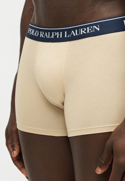 Άνδρας με μπεζ boxer Polo Ralph Lauren με ναυτικό μπλε μέση και λευκό λογότυπο, στέκεται σε ουδέτερο φόντο.