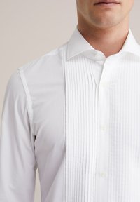 Balto smokingu krekls ar krokotu priekšu, izplestu apkakli un pogu aizdari. Izgatavots no gludas auduma ar klasisku piegriezumu.