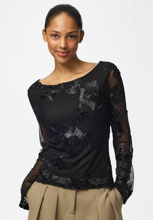 Femme portant un haut noir transparent à manches longues avec broderies florales et un pantalon plissé beige, souriante, les mains dans les poches.