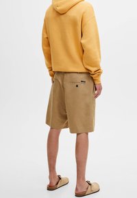 Sudadera amarilla, pantalones cortos beige con bolsillos traseros y zapatos de slip-on en tono marrón claro. La tela parece suave, con un corte relajado y mínimos detalles.