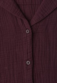Gros plan sur une chemise texturée couleur grenat avec boutons, col plié et deux boutons ronds visibles.