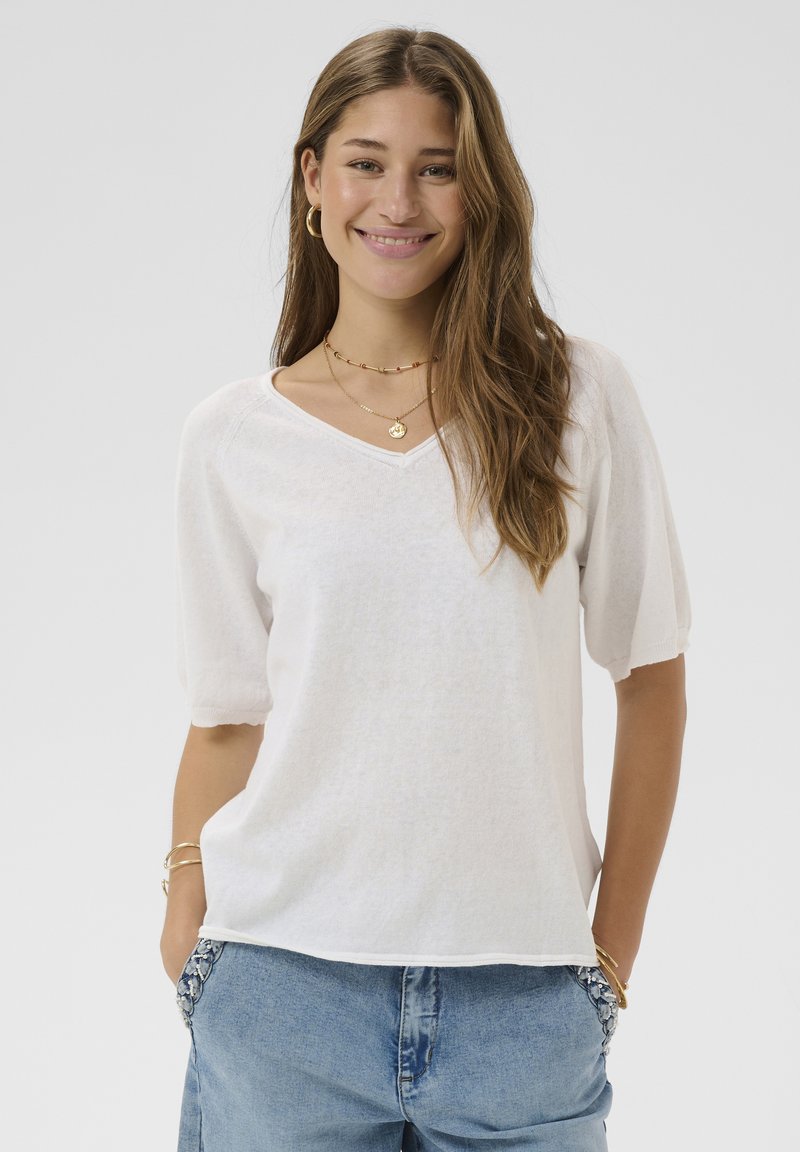 Top bianco a scollo a V realizzato in tessuto leggero con maniche corte a sbuffo, abbinato a jeans di denim blu con cuciture decorative sulle tasche.