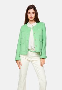 Mintgrüne Jacke mit strukturierter Oberfläche, runder Ausschnitt, frontale Taschen und silberne Knöpfe, kombiniert mit weißen Hosen und einem passenden Gürtel.