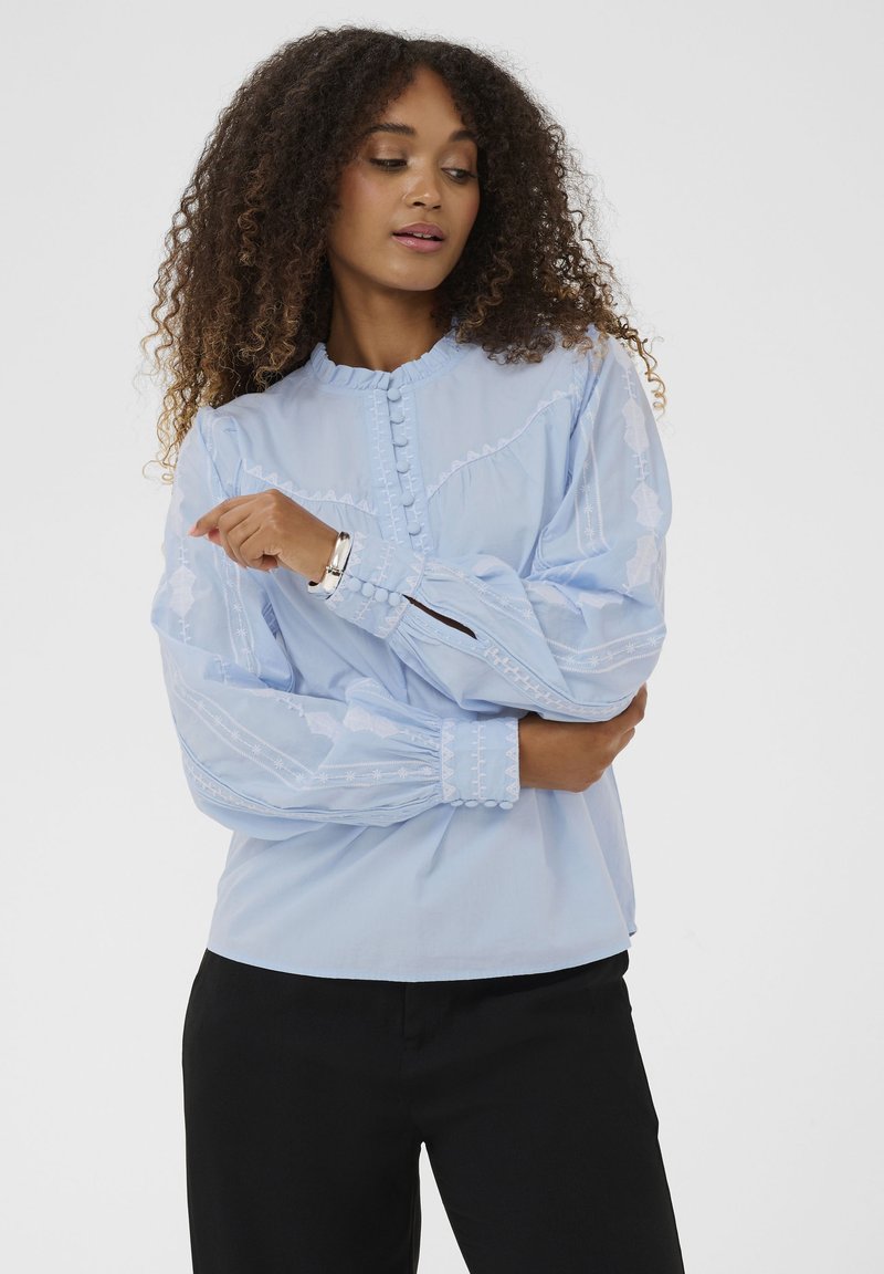 Blouse bleu clair avec des détails brodés, col rond, fermeture par boutons, et manches volumineuses. Portée avec un pantalon noir.
