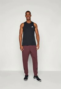 Μαύρο αμάνικο αθλητικό τοπ με λογότυπο Adidas, συνδυασμένο με μπορντό φόρμες που διαθέτουν μαύρες λεπτομέρειες και γραμμές, φορεμένο με μαύρα αθλητικά παπούτσια.