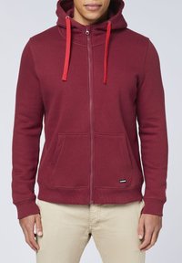 Bordeauxfarbener Zip-Hoodie, figurbetontes Design, aus Baumwollmischung, mit roten Kordeln und Fronttaschen. Auffälliges Markenetikett am Saum.