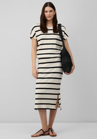 Gestreiftes Kleid in Creme und Schwarz, kurze Ärmel, tailliertes Design mit seitlichen Schlitzen. Getragen mit schwarzen Riemensandalen und einer gehäkelten Tasche.