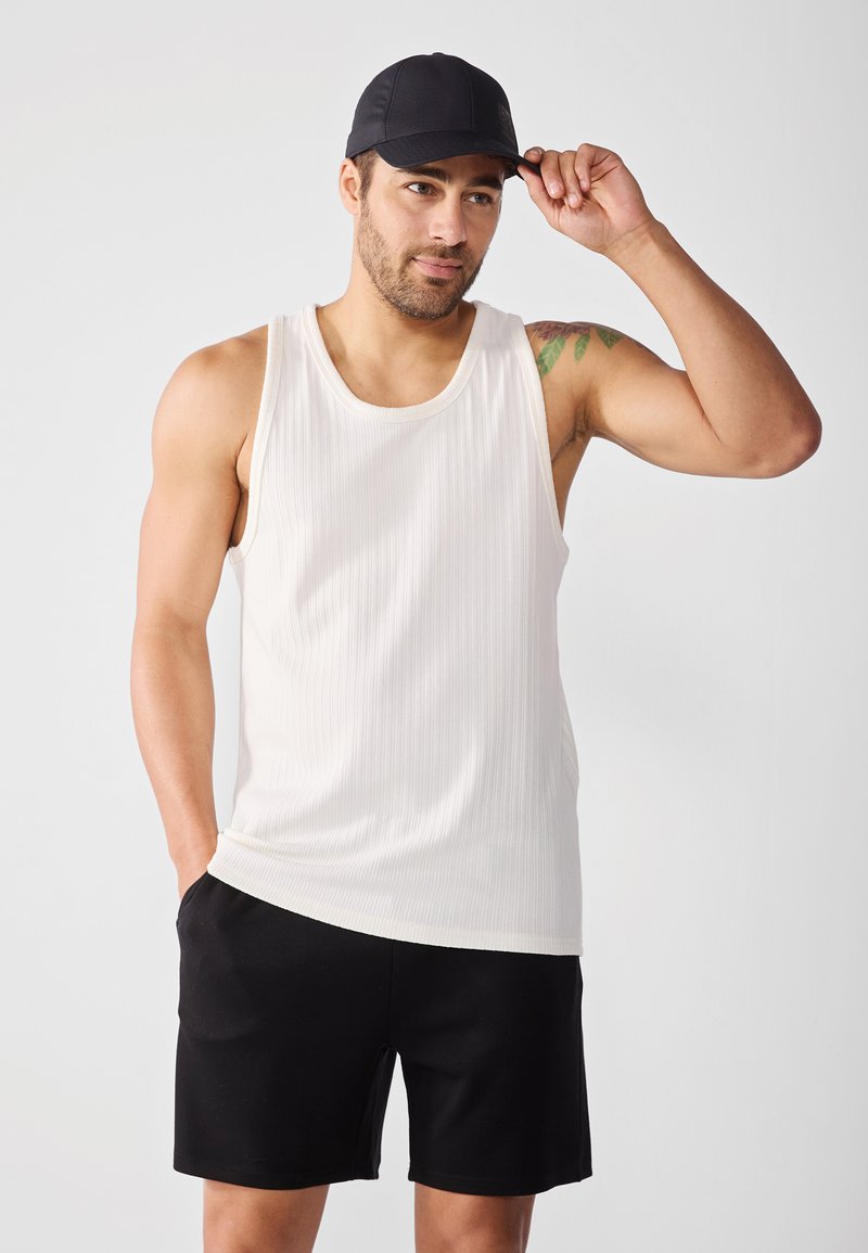 Kerge valge ribakujuline tank top, mida on kombineeritud mustade lühikestega. Mudel kannab musta mütsi, tal on lühike habet ja tätoveeringud käel.