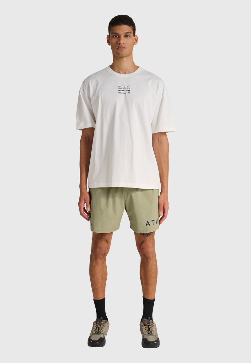Junger Mann steht frontal, trägt ein weißes übergroßes T-Shirt mit Text auf der Brust, hellgrüne Shorts mit schwarzen Buchstaben, schwarze Socken und beige Turnschuhe.