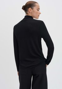 Schwarzes Langarmoberteil mit hohem Kragen, aus glattem, dehnbarem Stoff und einem lässigen Schnitt, in maßgeschneiderte schwarze Hose gesteckt.