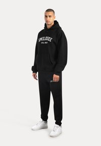 Czarna, oversize bluza z białym logo "SMILODON ATHL WEAR", w zestawie z pasującymi czarnymi spodniami dresowymi z małym logo, noszona w towarzystwie białych sneakersów.