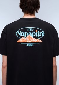 Sort kortærmet t-shirt med grafisk print på ryggen, der viser bjerge i orange, samt lys blå tekst med teksten "Napapijri" og "1987."