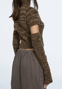 Top corto lavorato a maglia marrone con maniche corte e maniche allungate. Presenta un collo alto e un motivo testurizzato. Indossato con pantaloni grigi a vita elastica.