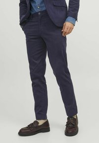 Pantalone od kordura u tamnoplavoj boji, uskog kroja s karakterističnim teksturiranim utorima. Upareni s tamnosmešnim cipelama s naglaskom na mašnu.