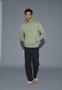 Lichtgroene hoodie met een voorzak en een trekkoord, gecombineerd met zwarte cargobroek en beige sneakers. Eenvoudig ontwerp met subtiele branding.