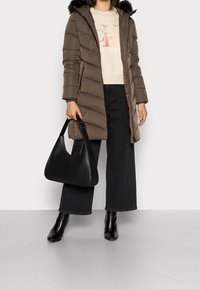 Kurtka puchowa w brązowym kolorze z futrzanym kapturem, połączona z kremowym swetrem, szerokimi czarnymi dżinsami oraz czarną torbą na ramię. Czarne botki za kostkę.