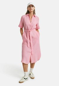 Abito camicia di lino rosa con maniche corte, chiusura a bottoni davanti, cintura in vita, tasche laterali e orlo fino al ginocchio. Indossato con sneakers e calze.
