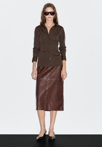 Braunes Hemd mit langen Ärmeln, kombiniert mit einem braunen Leder-Midi-Rock und schwarzen spitzen Ballerinas. Das Model trägt dunkle Sonnenbrillen.