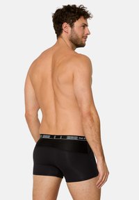 Man med lockigt hår, svagt v�änd åt sidan, iförd svarta boxerkalsonger med märkesresår mot vit bakgrund.